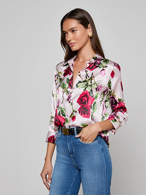 A woman with long brown hair wears the Dani Silk Blouse—a white, button-down top with a red rose print—paired with blue jeans, statement gold earrings, and a black belt. She stands against a plain light gray background, looking to the side.