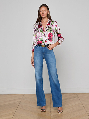 A woman with long brown hair poses indoors against a plain white wall, wearing the Dani Silk Blouse with a rose print, tucked into high-waisted blue jeans accessorized with a black belt and heeled sandals.
