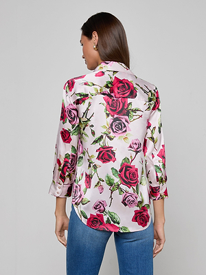 A woman with long brown hair, seen from behind, is wearing the Dani Silk Blouse with a red and pink rose print and blue jeans. The blouse features long sleeves, a relaxed fit, and button-down design in white silk.