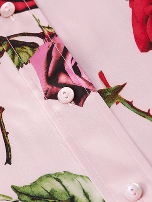 Close-up of the Dani Silk Blouse in light pink, showcasing red and purple rose prints with green leaves and stems, and detailed with white buttons along a visible seam.