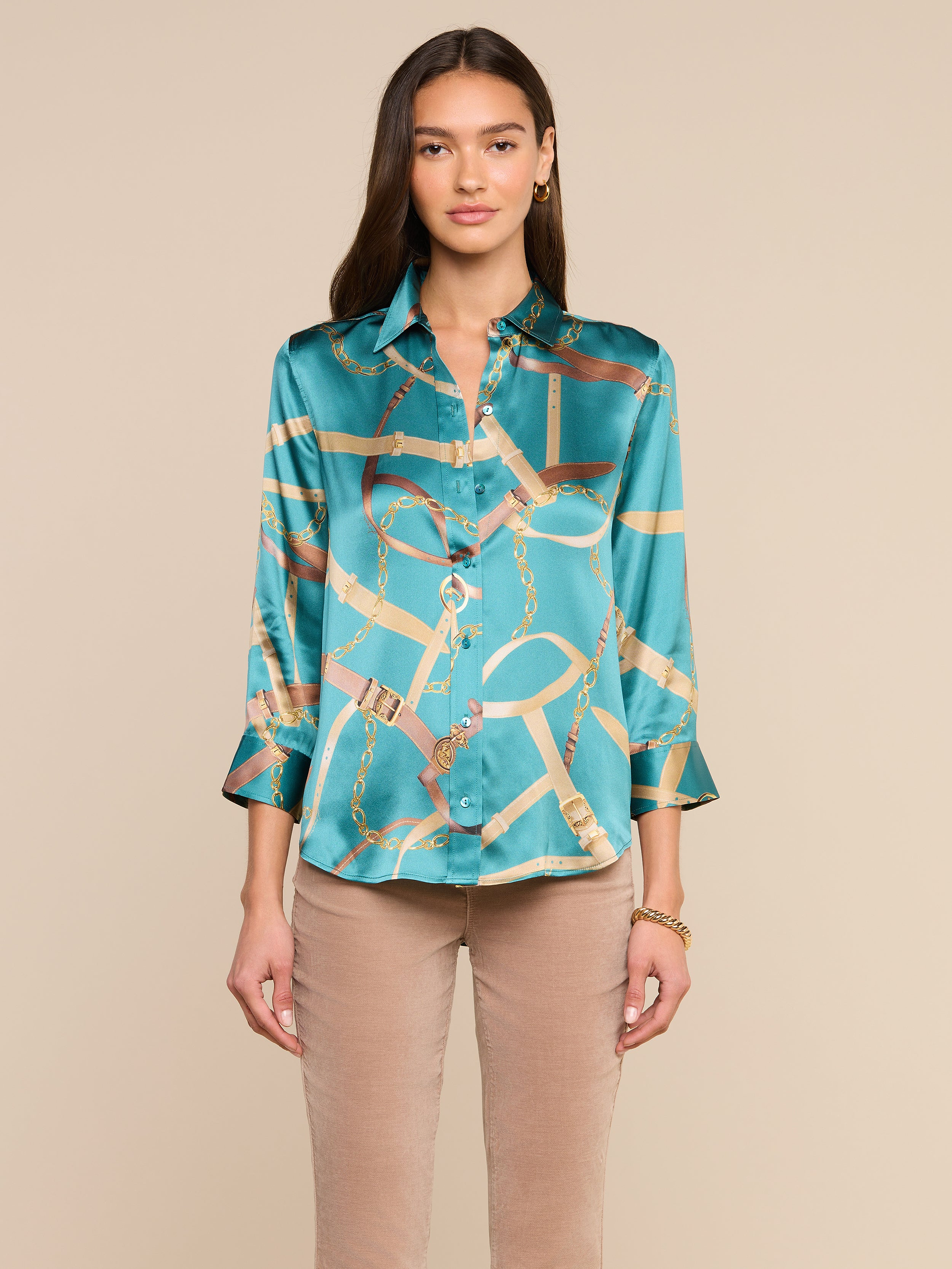 A woman stands facing forward, wearing the Dani Silk Blouse in teal, featuring a gold chain and belt pattern, paired with beige pants. Her long hair is worn loose, and she has a neutral expression against a plain background.