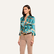 A woman wearing the Dani Silk Blouse in teal with a chain print, paired with tan pants and a brown belt, stands against a plain light background, looking slightly to the side.