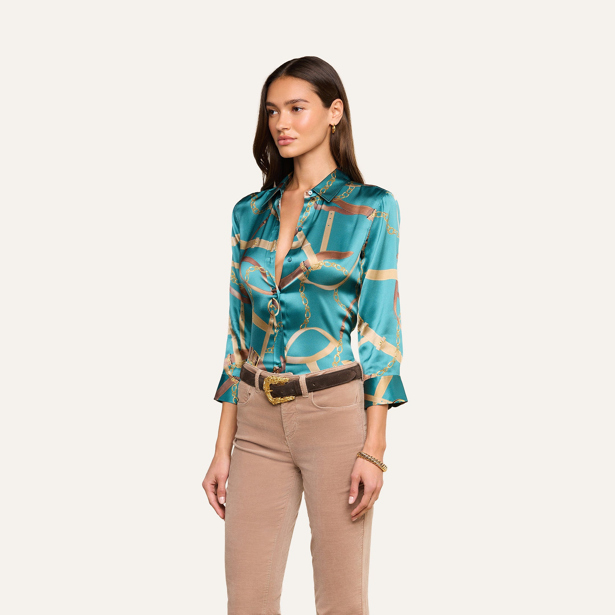 A woman wearing the Dani Silk Blouse in teal with a chain print, paired with tan pants and a brown belt, stands against a plain light background, looking slightly to the side.
