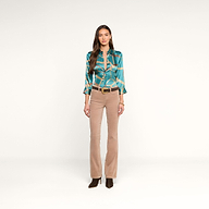 A woman stands facing forward against a plain white background, wearing the Dani Silk Blouse in a teal pattern, paired with beige pants, a belt, and dark heeled shoes.