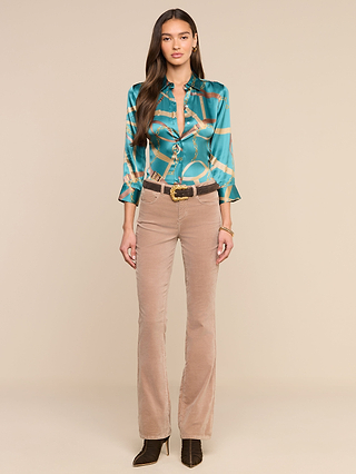 A woman stands facing forward against a plain beige background, wearing the Dani Silk Blouse in a teal pattern styled as a button-down, paired with tan high-waisted pants, a brown belt featuring a gold buckle, and heeled boots.