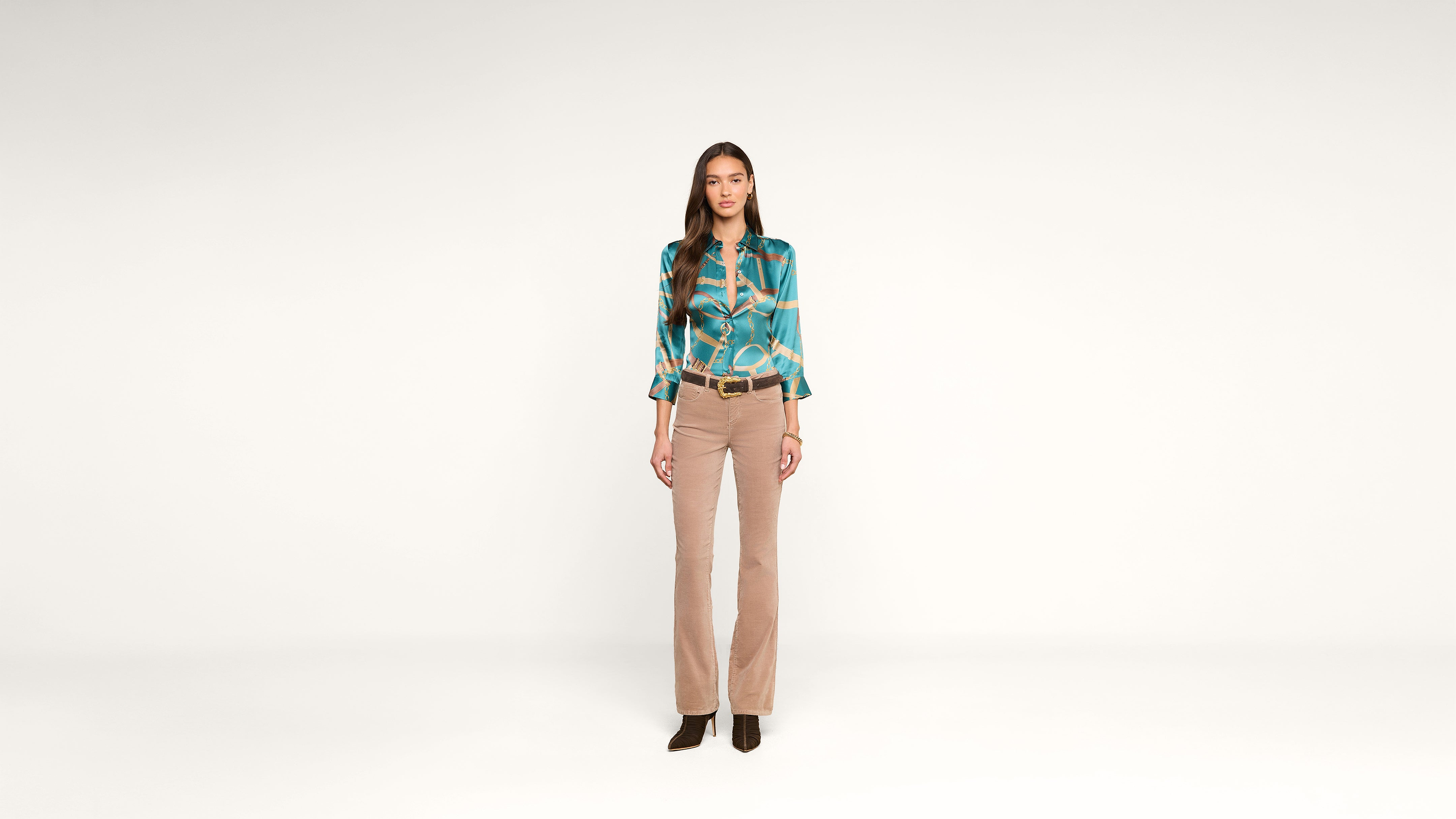 A woman stands facing forward against a plain white background, wearing a teal patterned blouse, beige pants, and black shoes.