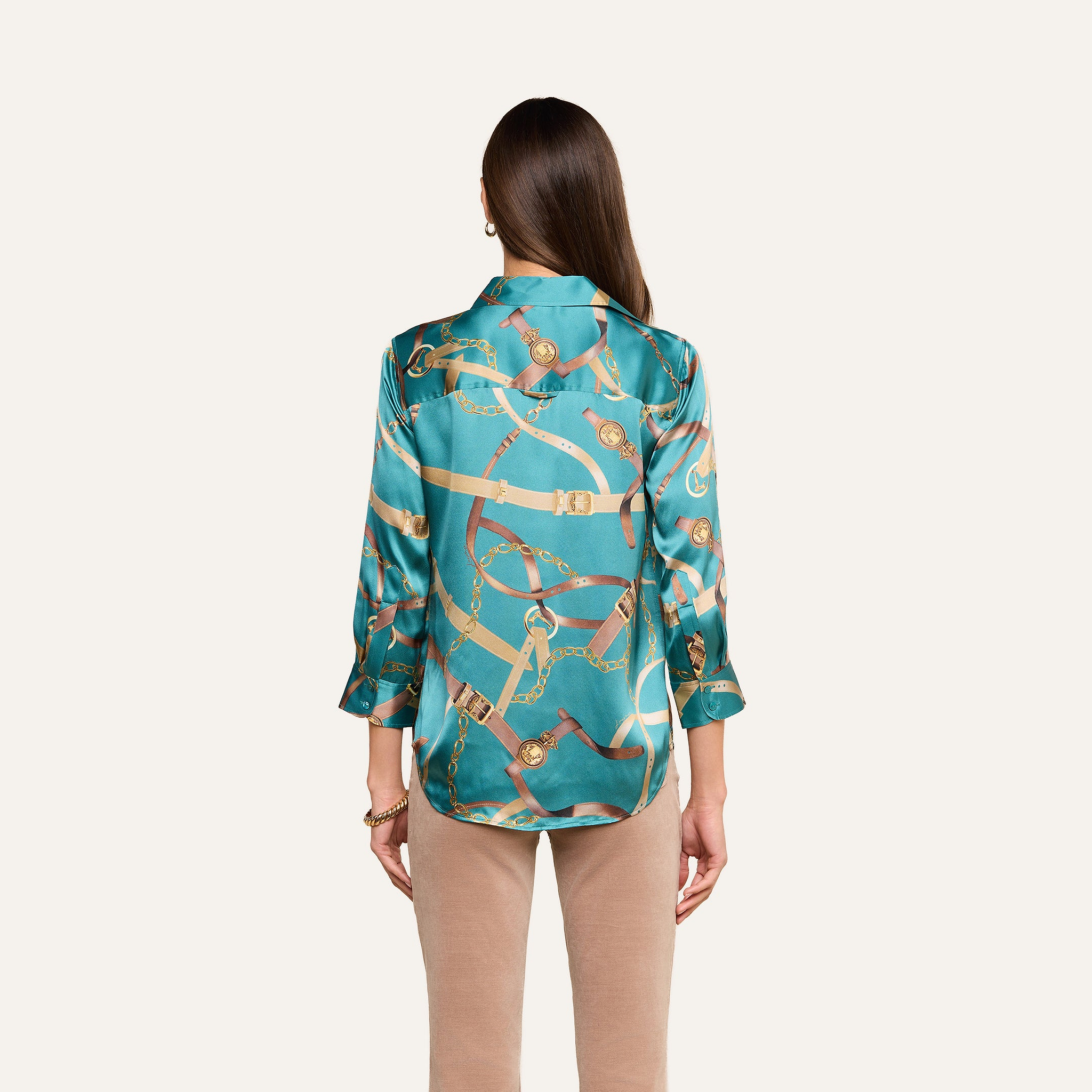 A woman stands facing away, wearing the Dani Silk Blouse featuring a teal belt print with gold and brown chain details, paired with beige pants, against a plain light background.