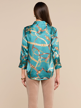 A woman with long brown hair, wearing the Dani Silk Blouse—a teal satin button-down adorned with elegant gold chain and belt motifs—and beige pants, stands with her back to the camera against a neutral background.