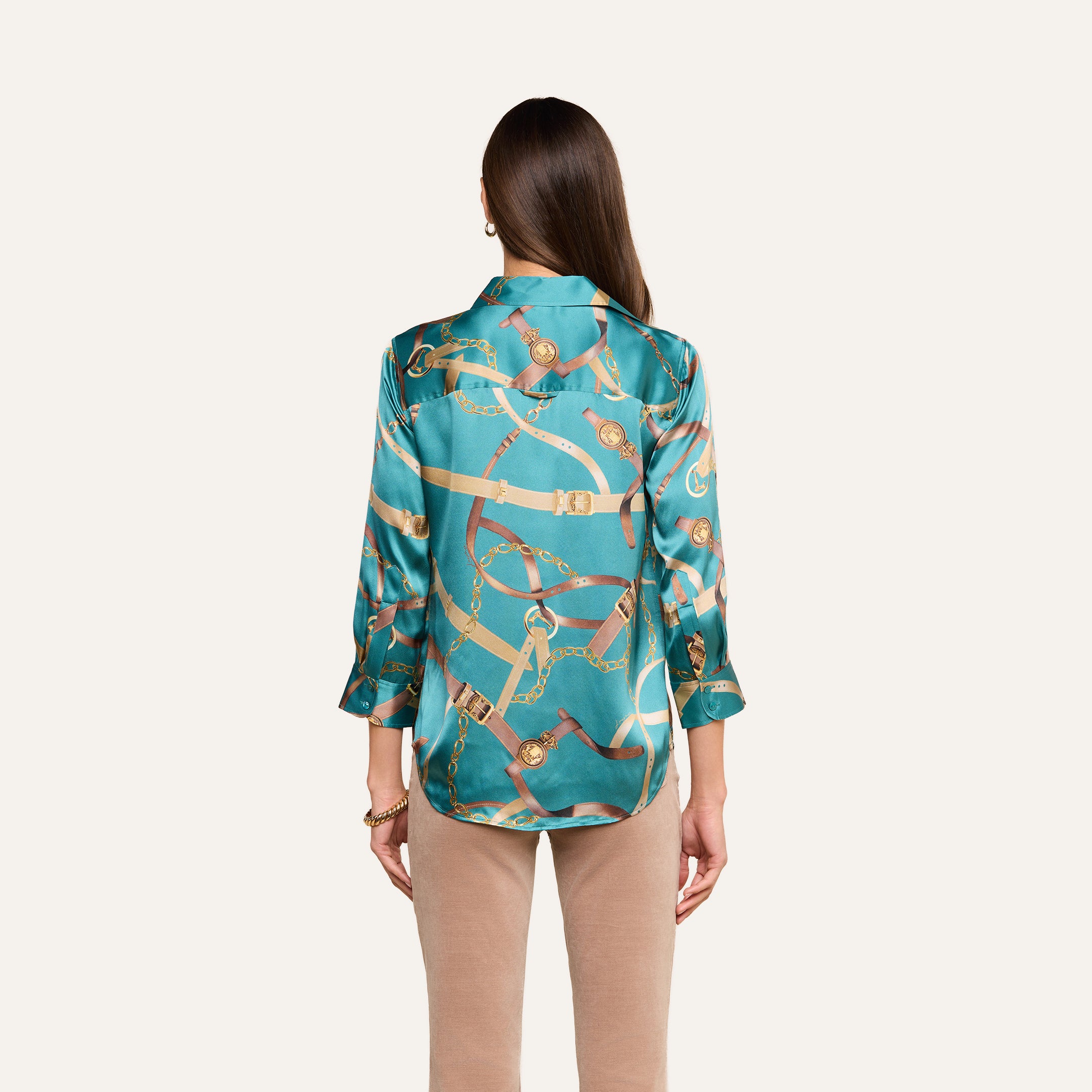 A woman stands facing away, wearing the Dani Silk Blouse featuring a teal belt print with gold and brown chain details, paired with beige pants, against a plain light background.