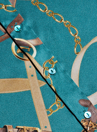 Close-up of the Dani Silk Blouse in teal, showcasing a gold chain and belt print pattern. The design features two pearly buttons and a diagonal seam, while the silky fabric captures the sophisticated style of a classic silk button-down.