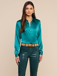 A woman wearing the Tyler Silk Blouse in teal paired with dark green pants stands facing forward with her hands by her sides against a plain light background.