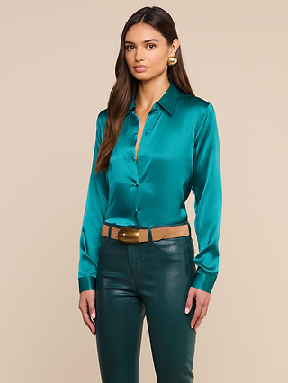 A woman with long brown hair wears the Tyler Silk Blouse in teal, paired with green leather pants, a tan belt featuring a gold buckle, and gold earrings. She stands against a plain beige background, looking calmly at the camera.