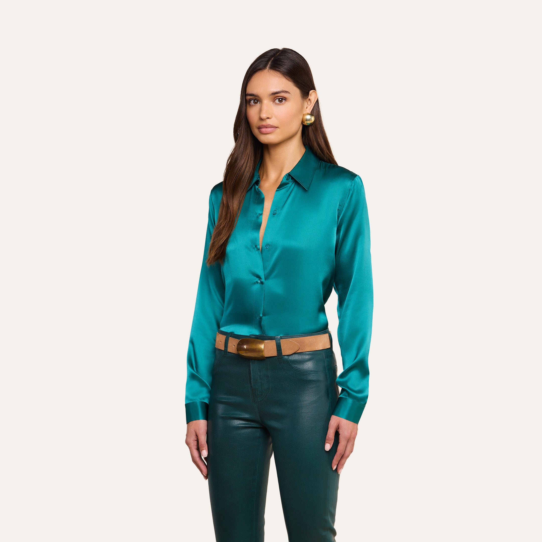 A woman with long brown hair models the Tyler Silk Blouse in teal, paired with dark green pants, a tan belt featuring a large buckle, and gold earrings, standing against a plain light background.