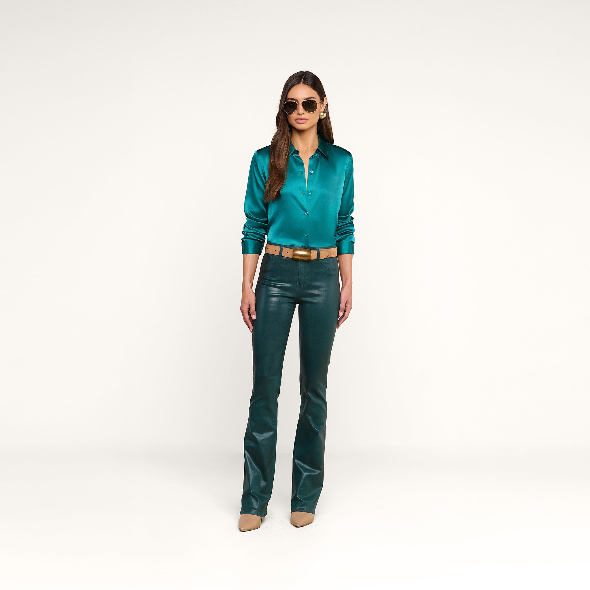 A woman wearing sunglasses, the Tyler Silk Blouse in teal, dark green leather pants, and beige heels stands against a plain white background.