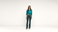 A woman wearing sunglasses, a teal blouse, teal leather pants, a tan belt, and beige shoes stands against a plain white background.