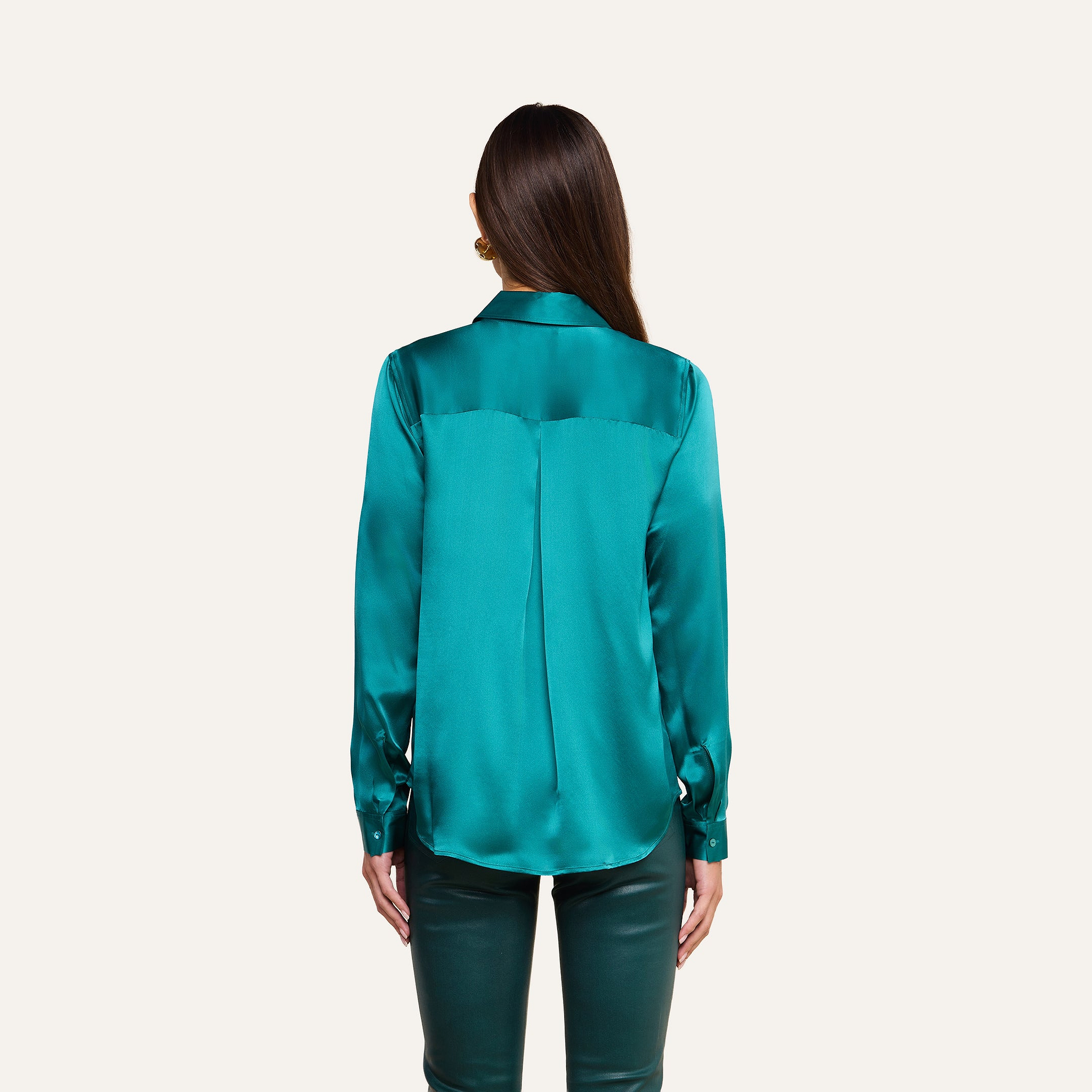A woman faces away against a plain light background, wearing the Tyler Silk Blouse in teal with matching teal pants. Her straight brown hair is loose, and her arms rest by her sides.