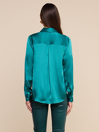 A person with long brown hair is shown from behind, wearing the Tyler Silk Blouse—a shiny teal button-down blouse with long sleeves—and matching teal pants against a beige background.