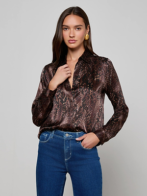 A woman with long dark hair wears the L'AGENCE Tyler Silk Blouse in a shiny snake print, tucked into high-waisted blue jeans, accessorized with gold earrings, standing against a plain light background.