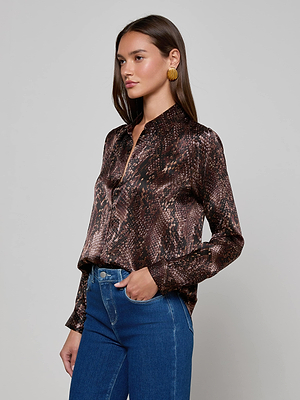 A woman with long brown hair wears the Tyler Silk Blouse by L'AGENCE—a snake print, collared, button-down top—paired with blue jeans and gold earrings. She stands sideways against a plain light background.