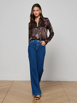 A woman with long hair poses indoors against a plain wall wearing the L'AGENCE Tyler Silk Blouse, blue high-waisted wide-leg jeans, and black open-toe heels, with one hand in her pocket.