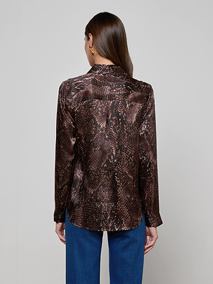 A woman with straight brown hair faces away, wearing the L'AGENCE Tyler Silk Blouse— a long-sleeve button-down in snake print—paired with blue jeans against a plain light background.