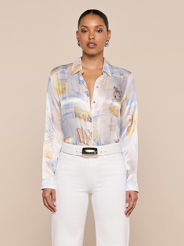 A woman stands against a plain background, wearing the L’AGENCE Tyler Silk Blouse in a pastel pattern, paired with high-waisted white pants and a black-buckled white belt.