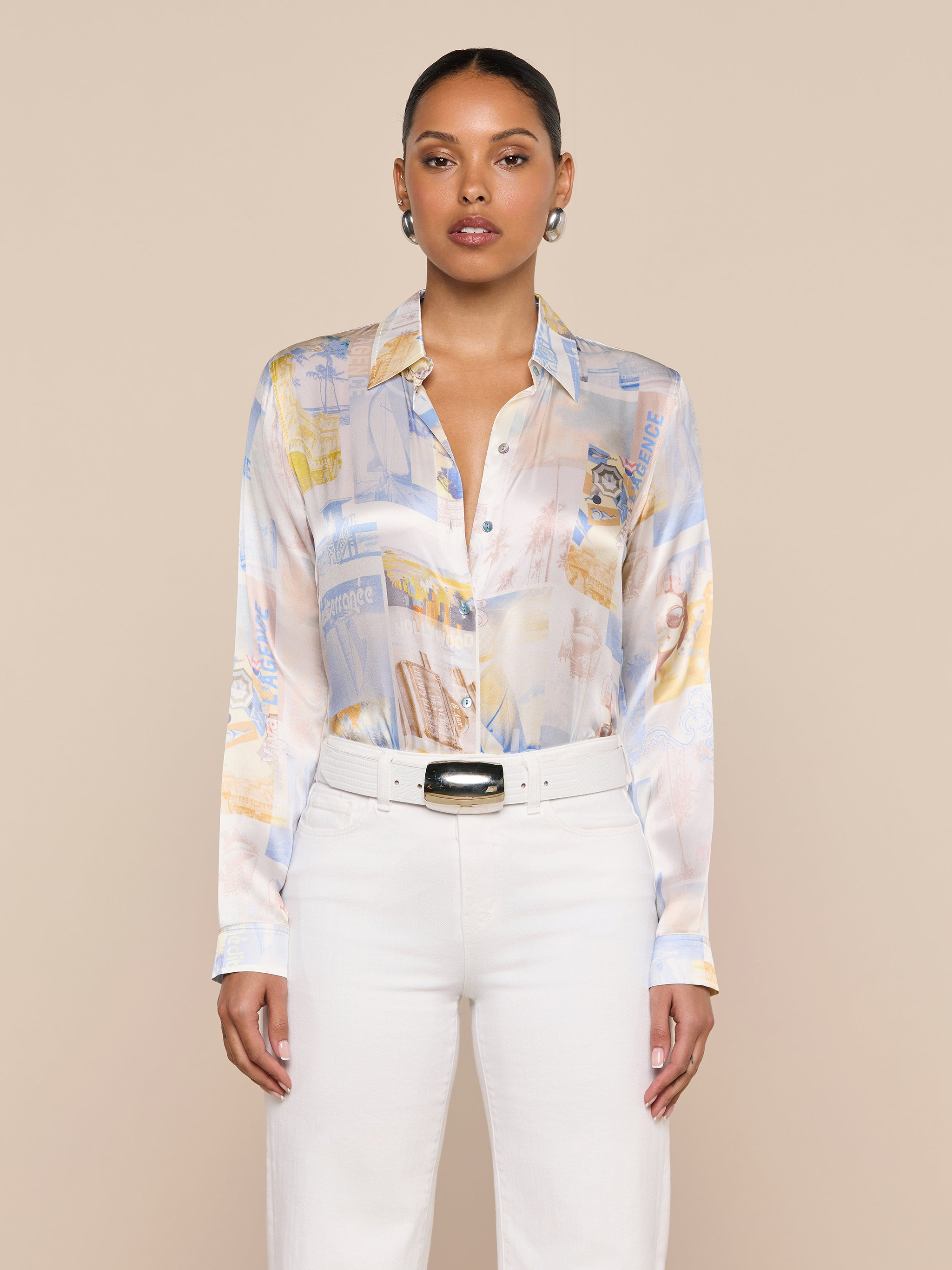 A woman stands against a plain background, wearing the L’AGENCE Tyler Silk Blouse in a pastel pattern, paired with high-waisted white pants and a black-buckled white belt.