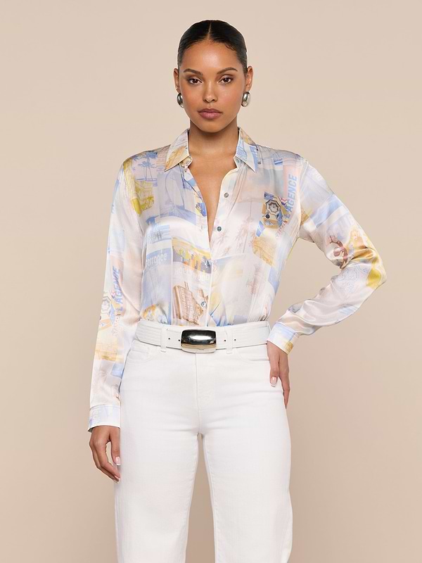 A woman stands facing forward, wearing the pastel patterned Tyler Silk Blouse by L’AGENCE, tucked into high-waisted white pants with a wide belt. Her hair is pulled back, and she accessorizes with earrings against a plain, light-colored background.