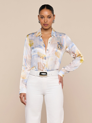 A woman with slicked-back hair confidently wears the L’AGENCE Tyler Silk Blouse, a patterned, long-sleeve button-down, paired with high-waisted white pants and a white belt against a plain beige background.