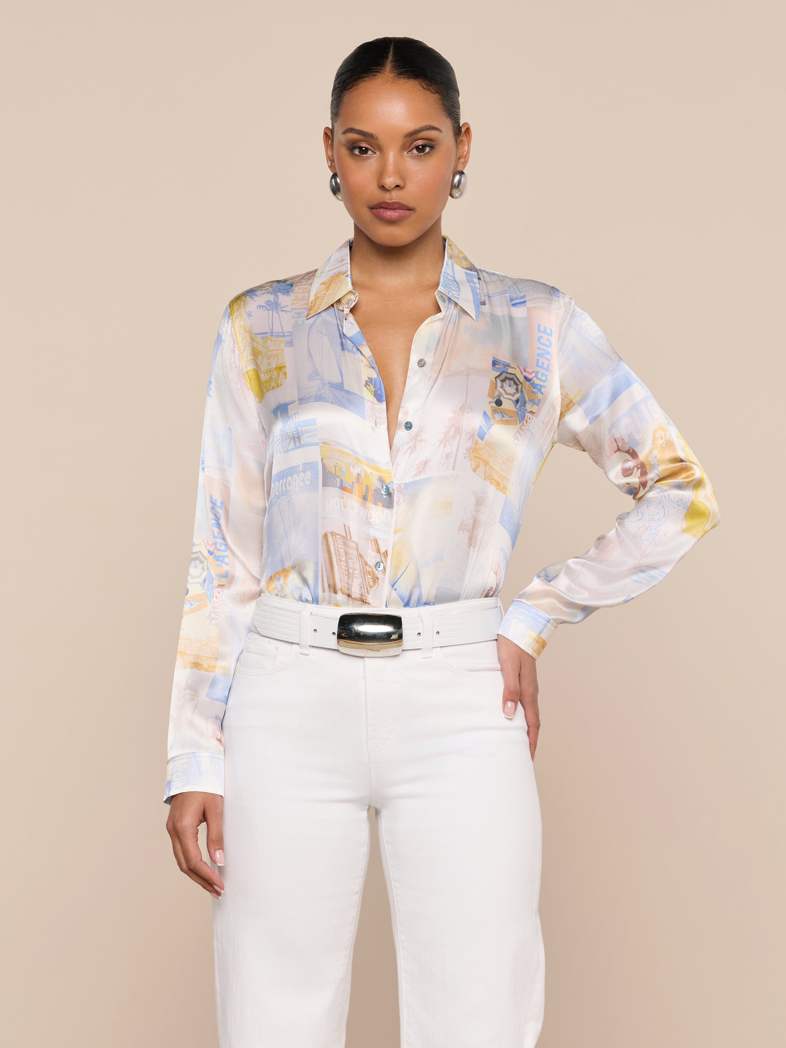 A woman stands facing forward, wearing the pastel patterned Tyler Silk Blouse by L’AGENCE, tucked into high-waisted white pants with a wide belt. Her hair is pulled back, and she accessorizes with earrings against a plain, light-colored background.
