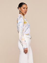 Woman standing sideways, wearing a L’AGENCE Tyler Silk Blouse in pastel colors with long sleeves, paired with white high-waisted pants and a white belt, against a plain light background.