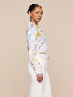 A woman stands in profile against a beige background, wearing the patterned Tyler Silk Blouse by L’AGENCE with long sleeves, high-waisted white pants, a white belt, large silver earrings, and her hair neatly pulled back.