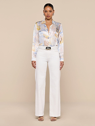 A woman stands facing forward against a plain white background, wearing the Tyler Silk Blouse—a pastel patterned silk button-down—tucked into high-waisted white pants, styled with sandals and a black belt.