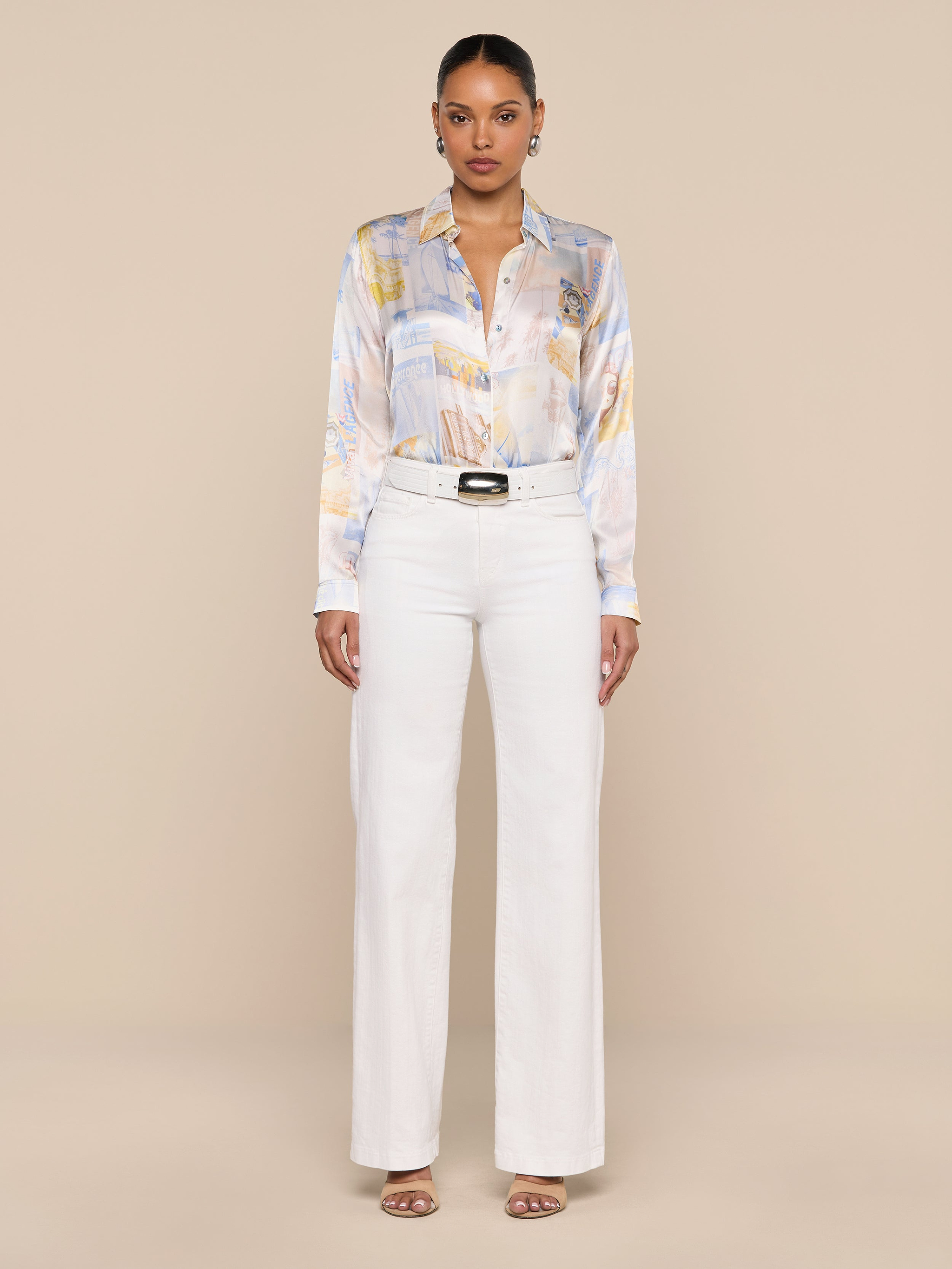 A woman stands facing forward against a plain white background, wearing the Tyler Silk Blouse—a pastel patterned silk button-down—tucked into high-waisted white pants, styled with sandals and a black belt.