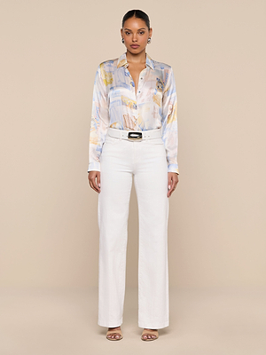A woman stands against a beige background, wearing the LAGENCE Tyler Silk Blouse in a pastel pattern with long sleeves, paired with white high-waisted wide-leg belted pants and open-toed heels. She faces forward with a neutral expression.
