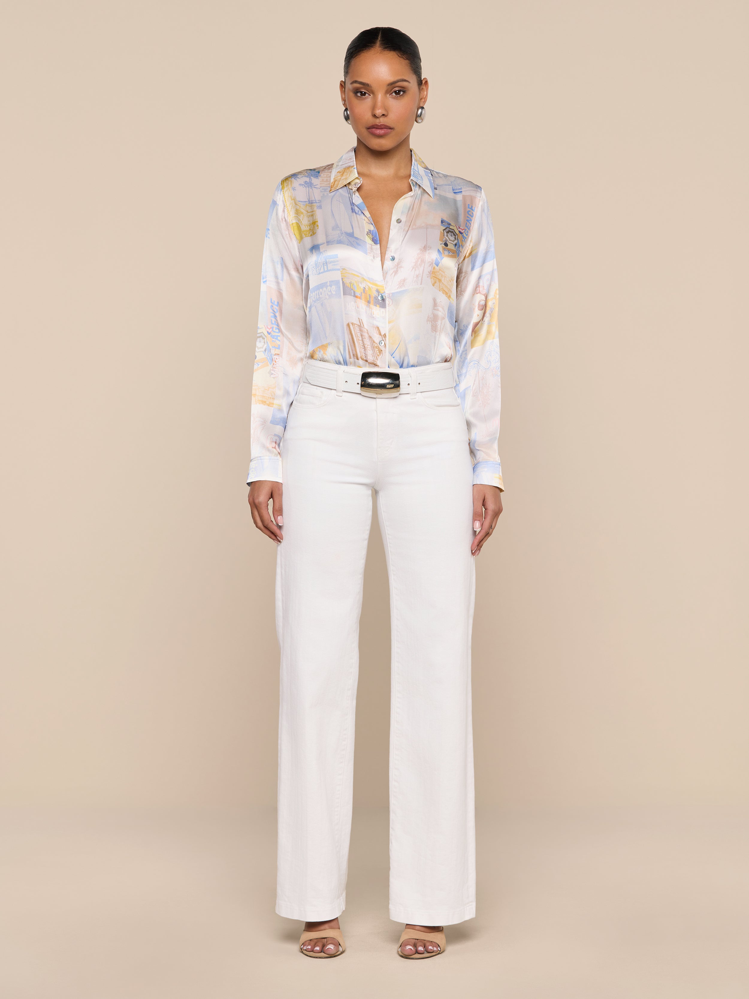 A woman stands facing forward against a plain white background, wearing the Tyler Silk Blouse—a pastel patterned silk button-down—tucked into high-waisted white pants, styled with sandals and a black belt.