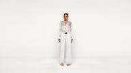 A woman stands facing forward against a plain white background, wearing a light-colored blouse with a subtle pattern, high-waisted white pants, and open-toe heels.