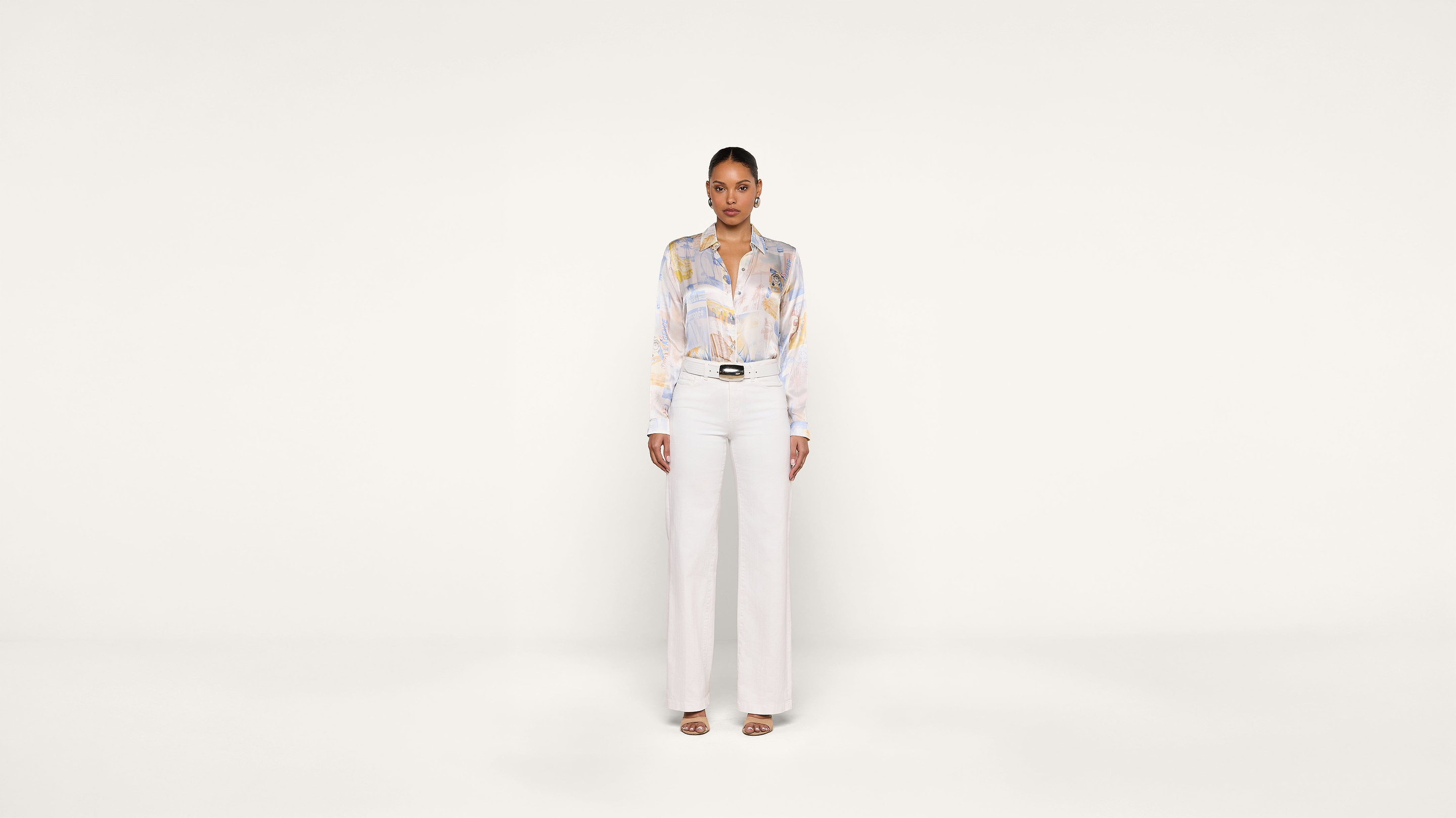A woman stands facing forward against a plain white background, wearing a light-colored blouse with a subtle pattern, high-waisted white pants, and open-toe heels.
