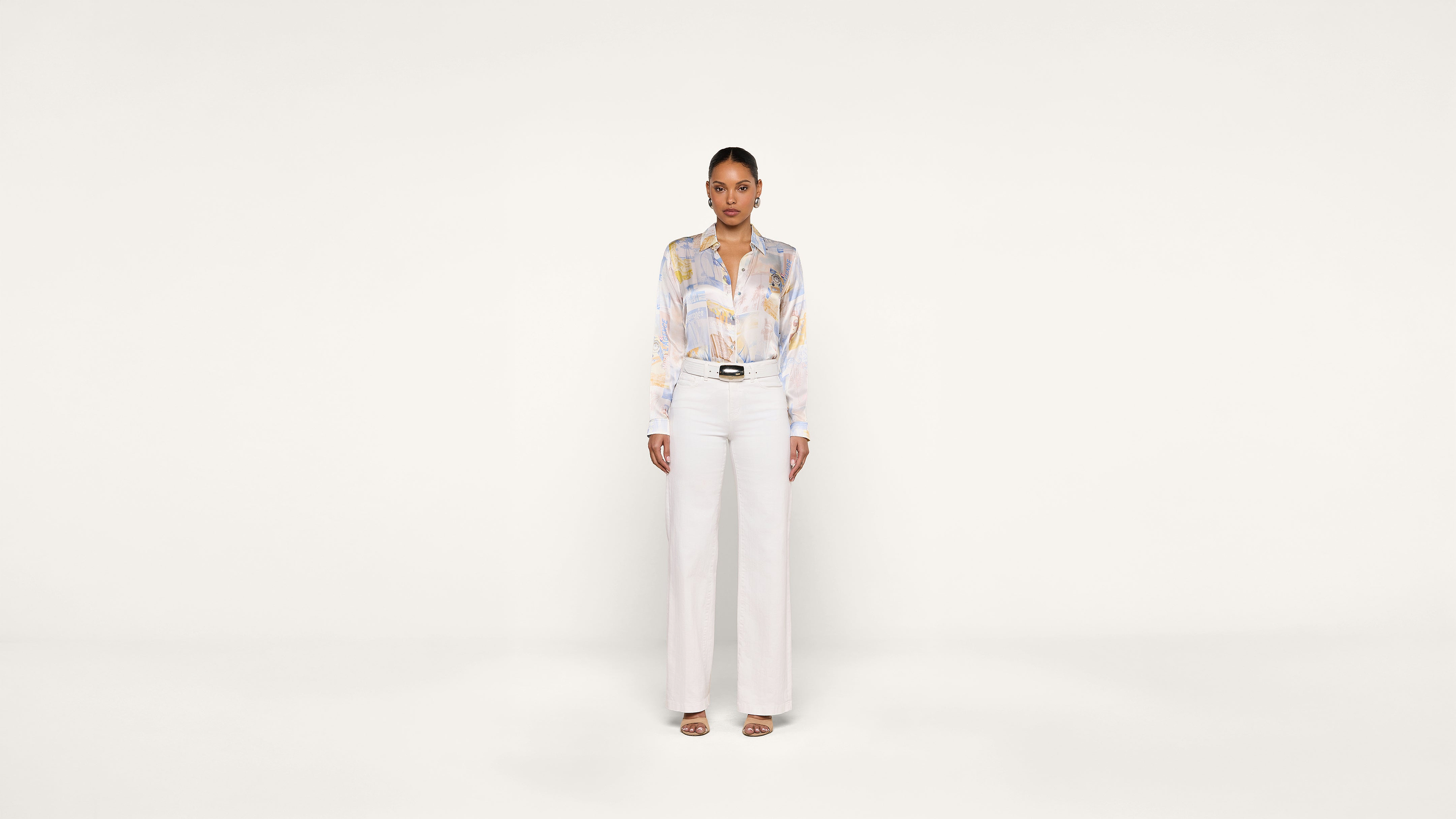 A woman stands facing forward against a plain white background, wearing a light-colored blouse with a subtle pattern, high-waisted white pants, and open-toe heels.