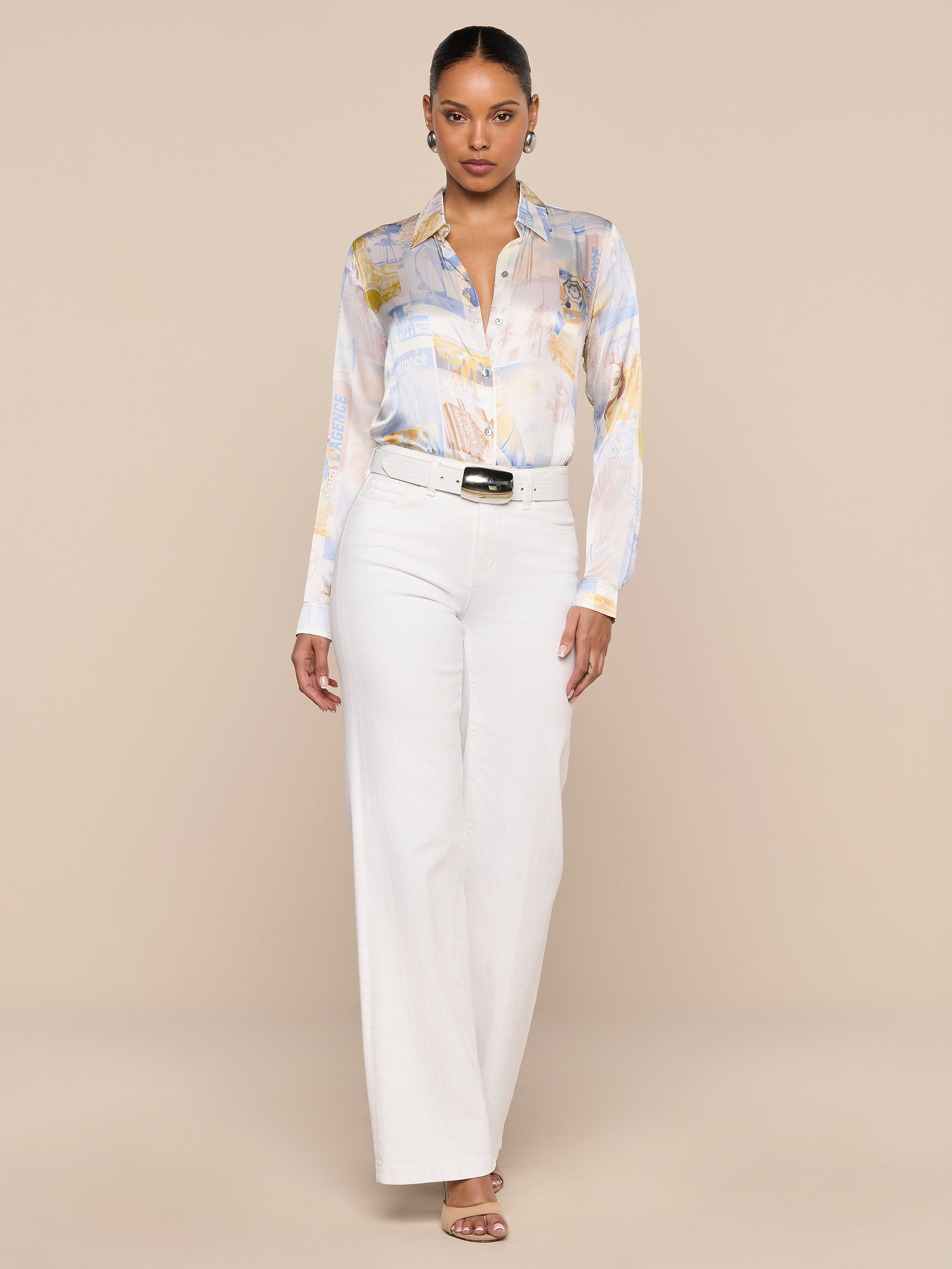 A woman stands facing forward, wearing the L’AGENCE Tyler Silk Blouse with a pastel pattern, tucked into high-waisted white wide-leg pants styled with a black belt and heeled sandals, against a plain white background.