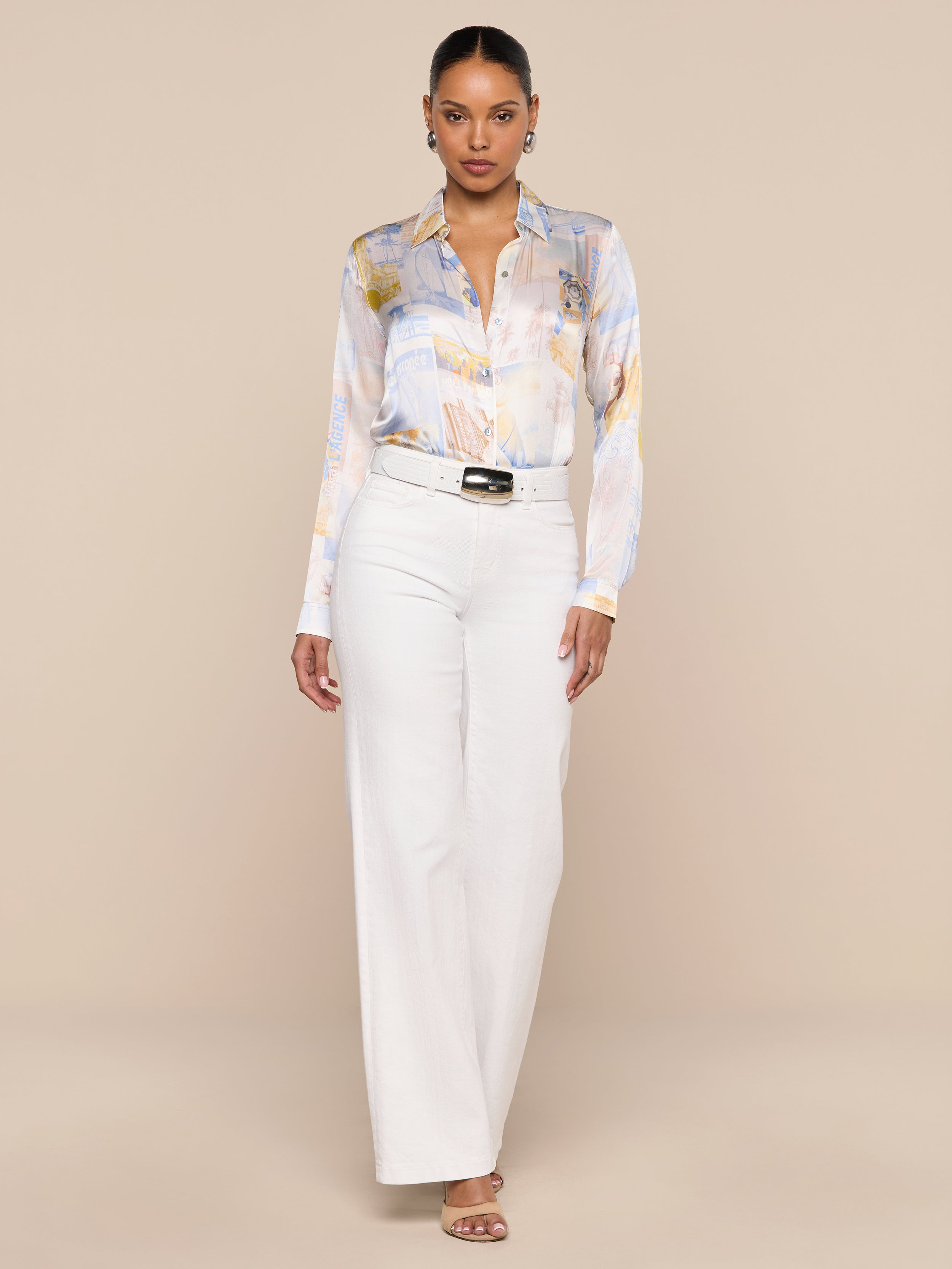 A woman stands facing forward, wearing the L’AGENCE Tyler Silk Blouse with a pastel pattern, tucked into high-waisted white wide-leg pants styled with a black belt and heeled sandals, against a plain white background.