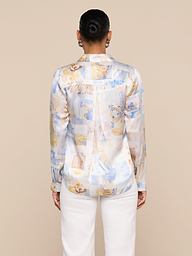 A person with their hair in a bun, wearing the Tyler Silk Blouse and white pants, stands facing away from the camera against a plain light background.