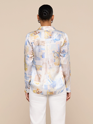 A person with hair in a neat bun wears the Tyler Silk Blouse, a light, patterned L’AGENCE button-down with long sleeves, paired with white pants, standing with their back to the camera against a plain beige background.