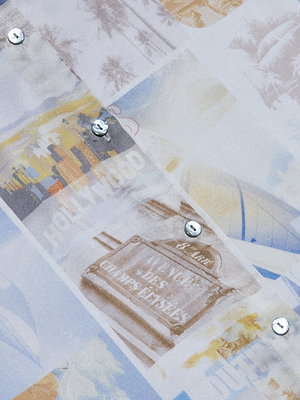 A close-up of the Tyler Silk Blouse fabric shows a faded collage of vintage travel postcards—Hollywood and beach scenes—with metal snap buttons on this silk button-down.