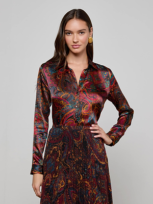 A woman with long brown hair wears the L'AGENCE Tyler Silk Blouse, a slim fit, long-sleeved, collared top featuring a medallion paisley print in red, blue, and gold. She stands with one hand on her hip against a plain light background.