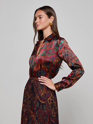 A woman with long brown hair wears the L'AGENCE Tyler Silk Blouse, styled slim fit with a medallion paisley print, paired with a matching skirt and gold round earrings, posing with one hand on her hip against a light gray background.