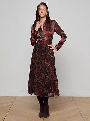 A woman stands indoors wearing the L'AGENCE Tyler Silk Blouse, featuring a colorful medallion paisley print in red, purple, and yellow. She pairs it with tall dark boots and poses with one hand touching her neck.