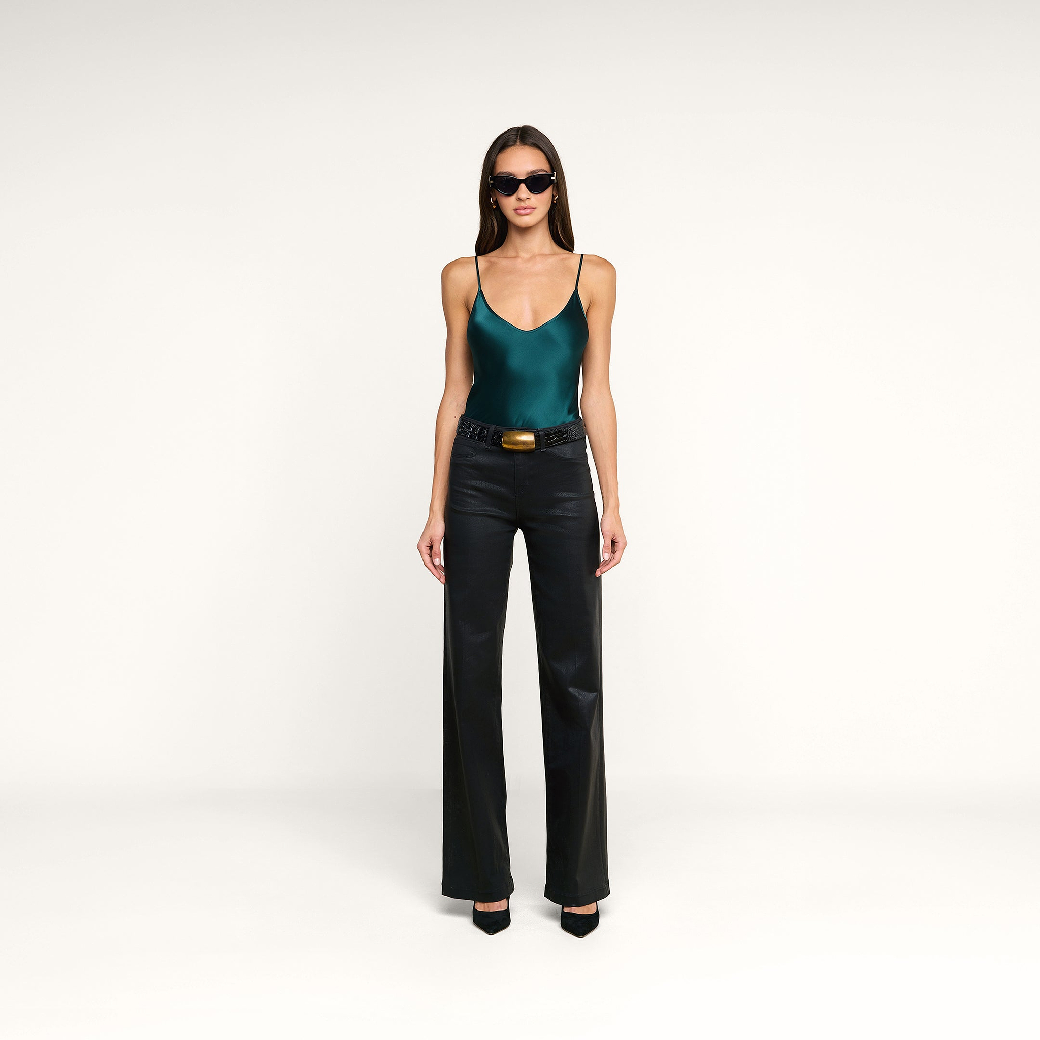 A woman wearing sunglasses, the Lexi Silk Camisole in teal with adjustable straps, black leather pants with a belt, and black heels stands against a plain white background.