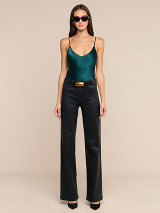 A woman stands against a beige background, wearing dark sunglasses, the Lexi Silk Camisole with adjustable straps in green, black leather wide-leg pants with a belt, and black heels.