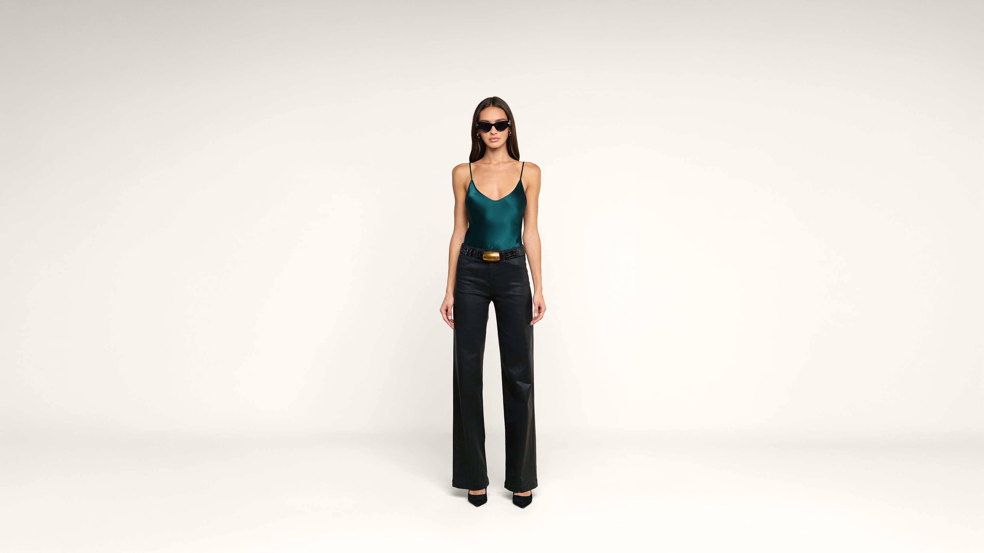 Woman in sunglasses wears a teal tank top, black flared pants, and black heels, standing against a plain white background.